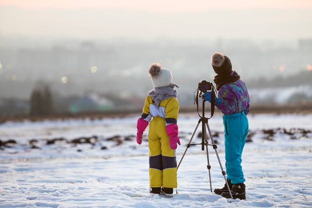 Deux enfants garçon et fille s'amusant à l'extérieur en hiver en jouant avec un appareil photo sur un trépied sur un terrain couvert de neige.