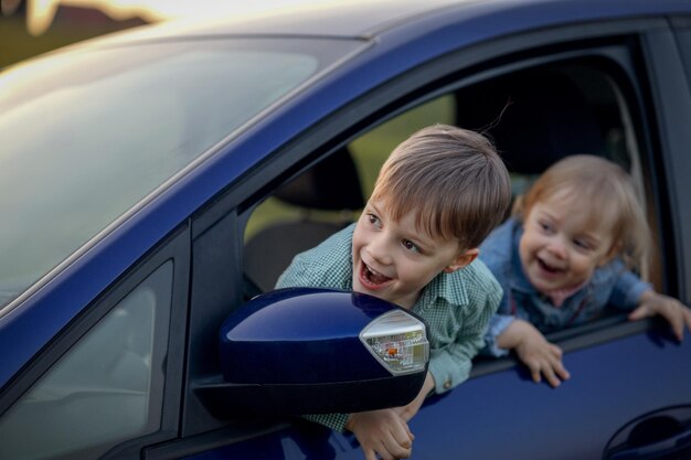 Deux enfants joyeux assis derrière le volant de la voiture de leur père se penchant par la fenêtre et riant