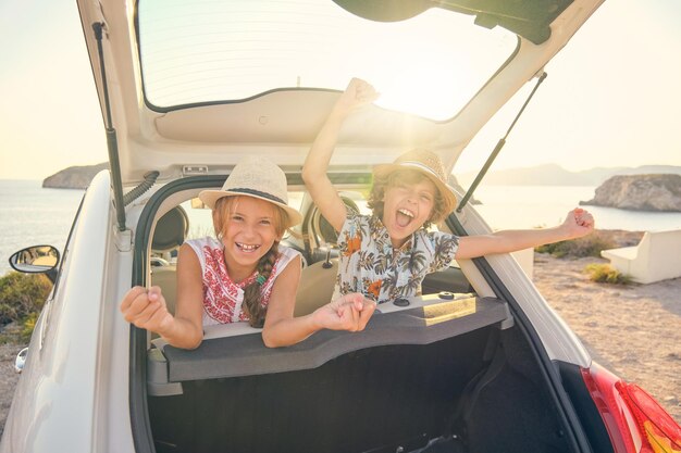 Deux enfants portant des chapeaux de paille assis sur les sièges arrière d'une voiture sortant la tête du coffre faisant des gestes de victoire avec les bras levés et une expression heureuse