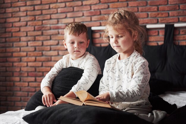 Deux enfants sont allongés sur un grand lit et lisent un livre intéressant. Ils sont vêtus du même pyjama