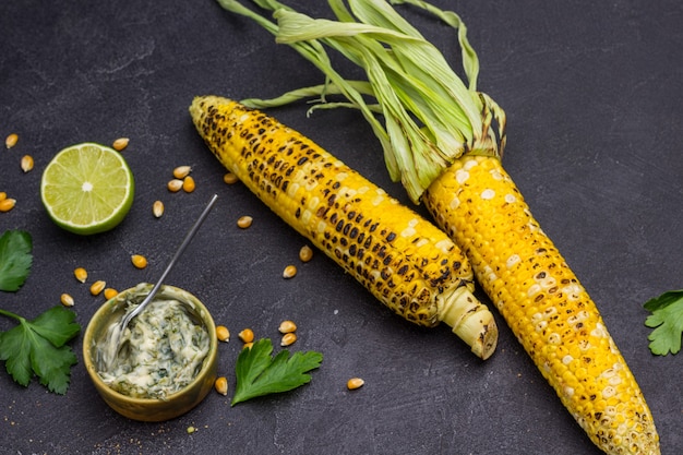 Deux épis de maïs grillés sur table. Sauce dans un bol et la moitié du citron vert. Fond noir. Vue de dessus