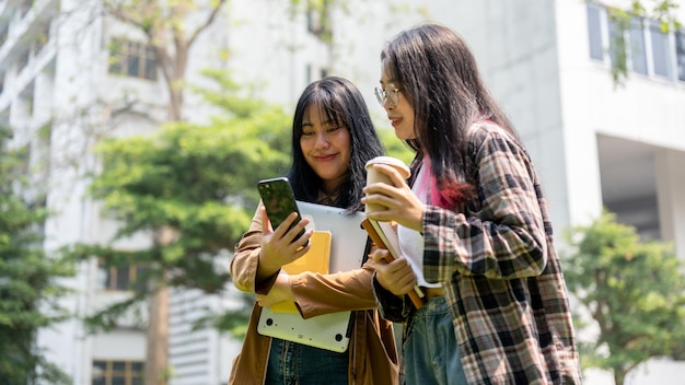 Deux étudiantes asiatiques se tiennent ensemble dans le parc du campus en train de regarder un smartphone.