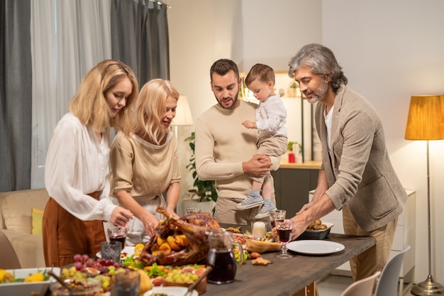 Deux femelles blondes et un homme aux cheveux gris mûr penché sur une table servie tout en mettant de la nourriture maison festive contre un jeune homme avec un petit fils