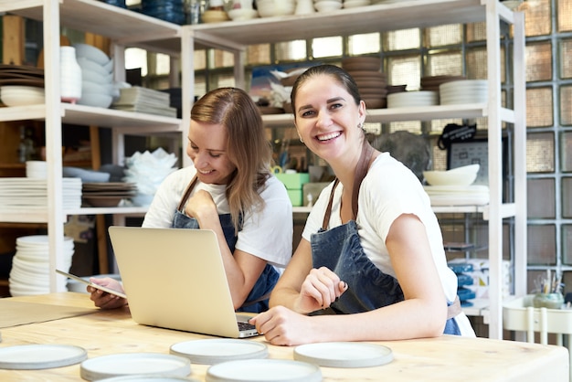 Deux femme entrepreneur souriante avec ordinateur portable dans l'atelier de travail artisanal.