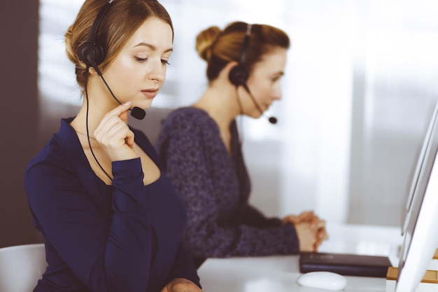 Deux Femmes D'affaires Ont Des Conversations Avec Les Clients Par Casques, Tout En étant Assises Au Bureau Dans Un Bureau Moderne. Groupe De Personnes Diverses Dans Un Centre D'appels. Télémarketing Et Service à La Clientèle.