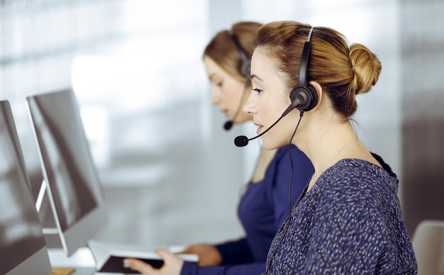 Deux femmes d'affaires ont des conversations avec les clients par casques, tout en étant assises au bureau dans un bureau moderne. Groupe de personnes diverses dans un centre d'appels. Télémarketing et service à la clientèle.