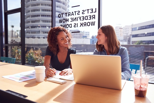 Deux femmes d'affaires avec ordinateur portable au bureau par fenêtre dans le bureau collaborant sur le projet ensemble