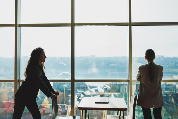 Deux femmes d'affaires se tiennent sur le fond d'une grande fenêtre dans un bureau moderne avec une vue panoramique sur la ville Les femmes d'affaires modernes travaillent à distance