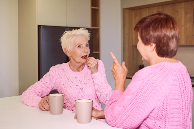 Deux femmes âgées élégantes en chandails roses assis avec des tasses à potins de cuisine moderne