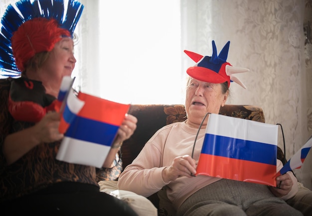 Deux femmes âgées enthousiastes regardant la télévision dans des accessoires russes tenant des drapeaux russes