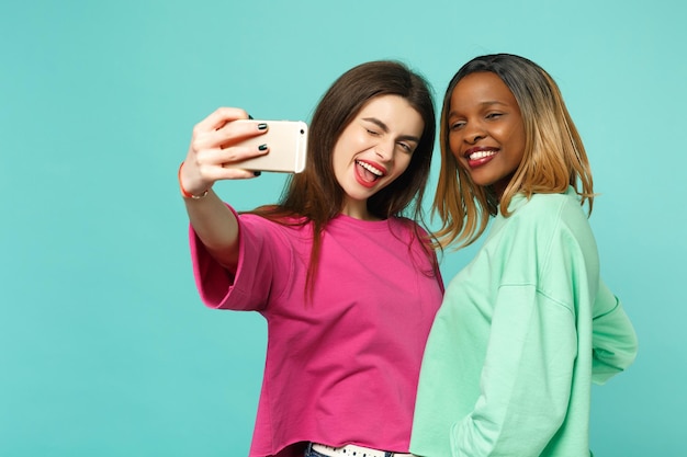 Deux femmes amies européennes et afro-américaines en vêtements verts roses faisant selfie tourné sur téléphone portable isolé sur fond de mur bleu, portrait en studio. Concept de style de vie des gens. Maquette de l'espace de copie.