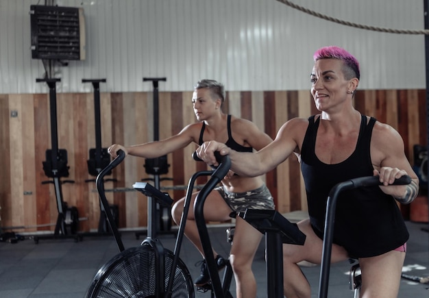 Deux femmes athlétiques musclées s'exerçant sur des vélos à air au gymnase. Entraînement fonctionnel cross class