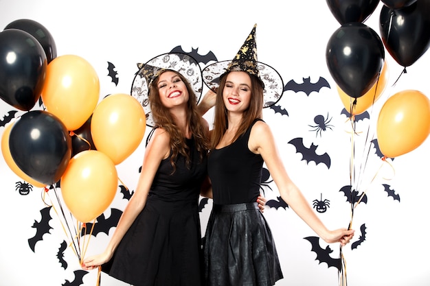 Deux femmes brunes en robes noires et chapeaux de sorcière tiennent des ballons noirs et oranges sur le fond du mur avec des chauves-souris. Fête d'Halloween .