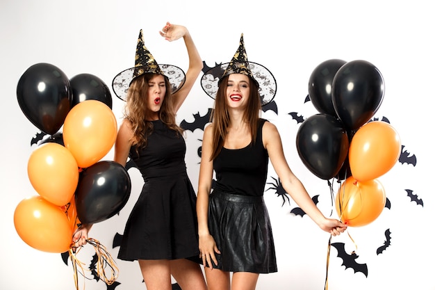 Deux femmes brunes en robes noires et chapeaux de sorcière tiennent des ballons noirs et oranges sur le fond du mur avec des chauves-souris. Fête d'Halloween .