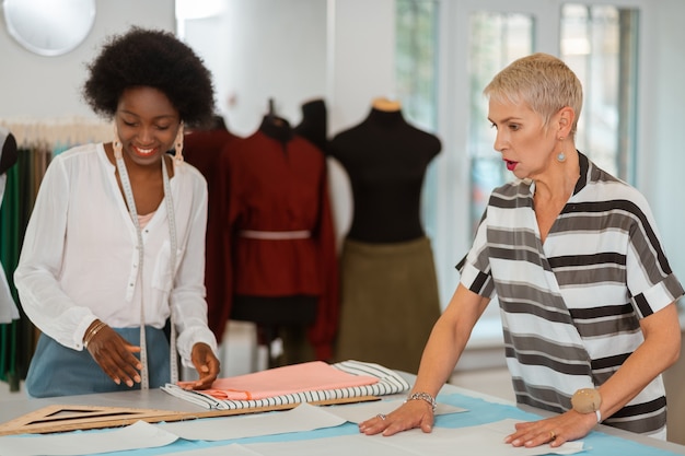 Photo deux femmes debout à côté du bureau et mettant les patrons en papier sur le tissu dans un atelier