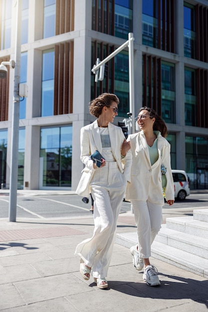 Deux femmes heureuses dans des tenues à la mode rient et marchent dehors sur fond de bâtiment moderne