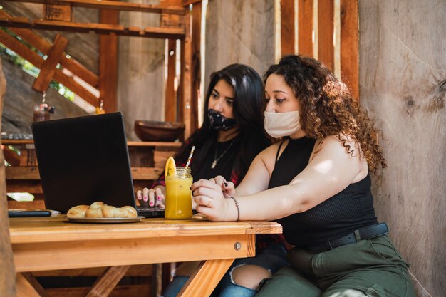 Deux femmes portant des masques de protection assis dans un bar travaillant avec un ordinateur portable - Femmes assises dans un bar en dégustant un jus.
