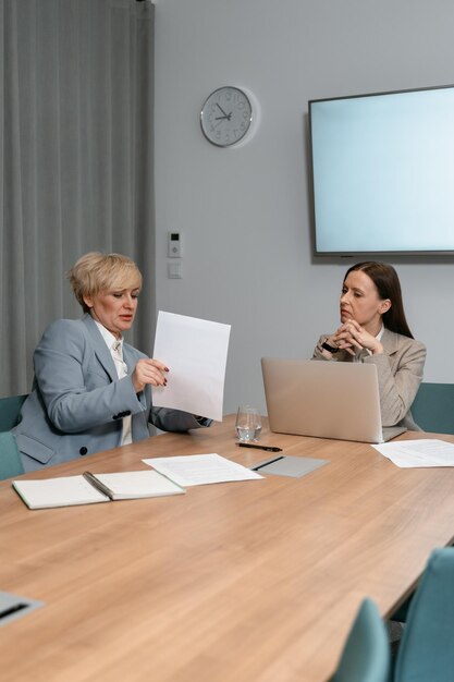 deux femmes sont assises à une table avec un ordinateur portable et une horloge sur le mur derrière elles