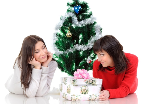 Deux femmes souriantes posant à côté d'un arbre de Noël et d'un cadeau. La fille tient les mains près du visage. Fond clair