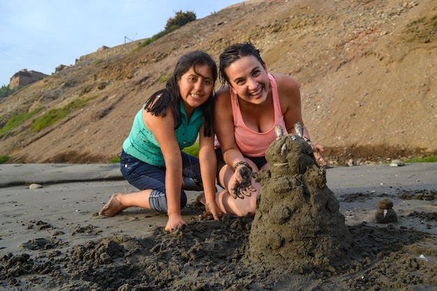 Deux femmes souriantes posent à côté d'une poupée de terre qu'elles ont montée au bord de la mer