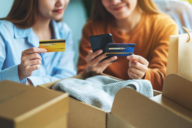 Deux femmes utilisant un téléphone portable et une carte de crédit pour faire des achats en ligne avec un sac à provisions et une boîte postale de vêtements sur la table