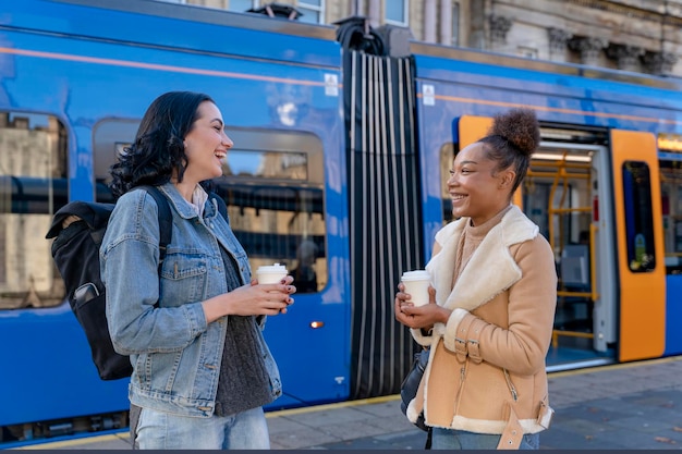 Deux femmes en veste en jean se parlent en riant en buvant du café et en attendant le tram