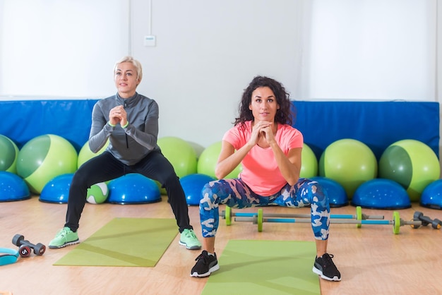 Deux femmes en vêtements de sport faisant des squats de poids corporel tout en s'entraînant à l'intérieur dans une salle de sport