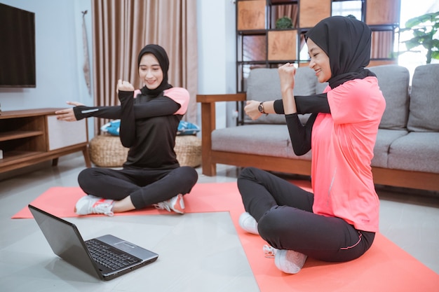 Deux femmes en vêtements de sport hijab assis les jambes croisées s'échauffant avec une main tenant l'autre main alors qu'une main est tirée sur le côté tout en faisant des activités ensemble à la maison