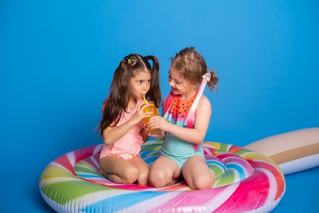 Deux fille d'enfant heureux en maillot de bain buvant du jus d'orange assis sur un matelas gonflable coloré.