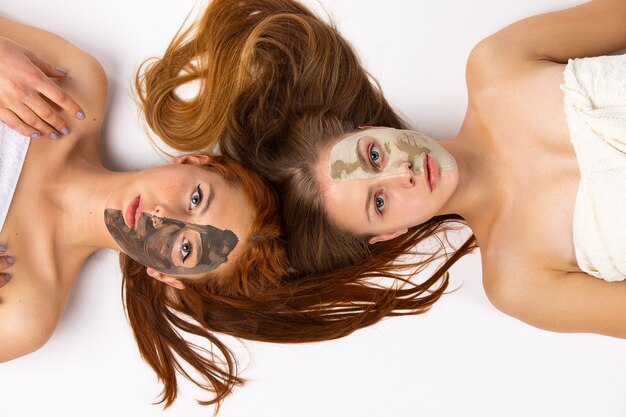 Deux filles, blondes et rousses, copines avec des masques différents sur la moitié du visage, allongées sur le sol enveloppées dans des serviettes. Peau saine et concept de beauté sur un mur blanc. Photo de haute qualité