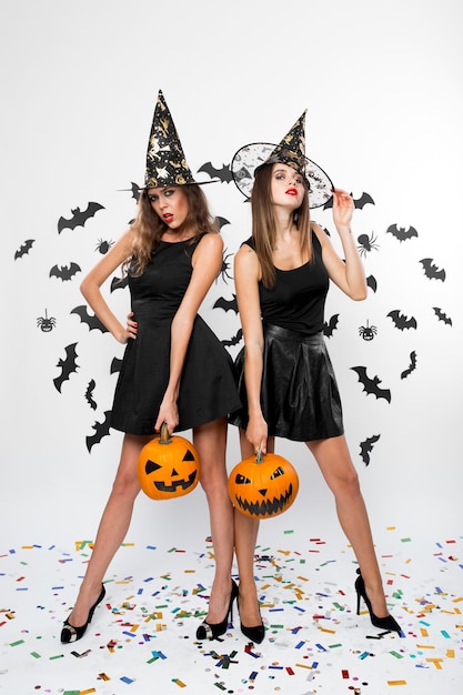Deux filles brunes portant des robes noires, des chapeaux de sorcière et des talons hauts tiennent des citrouilles d'halloween en arrière-plan avec des chauves-souris.