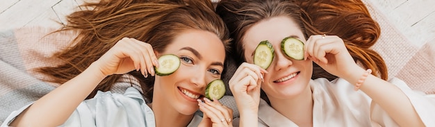 Deux filles fabriquent des masques de beauté pour le visage et les cheveux faits maison. Concombres pour la fraîcheur de la peau autour des yeux. Les femmes prennent soin d'une peau jeune. Les copines rient à la maison allongées sur le sol sur des oreillers.