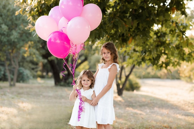 Deux filles heureuses portent des robes blanches tenant des ballons de décoration d'anniversaire dans le parc à l'extérieur sur la nature verdoyante Célébration Enfance