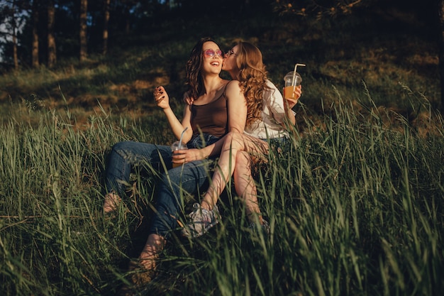 Deux filles heureuses s'amusent, s'assoient sur l'herbe, s'embrassent sur la joue, dans des lunettes de soleil, au coucher du soleil, expression faciale positive, en plein air