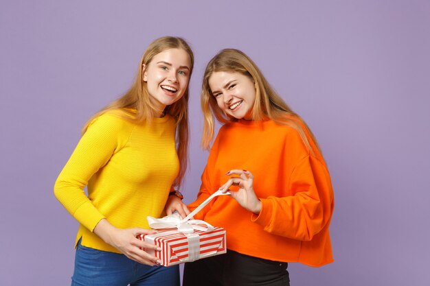 Deux Filles Jumelles Blondes Gaies Dans Des Vêtements Vifs Tiennent Une Boîte Cadeau à Rayures Rouges Avec Un Ruban Cadeau Isolé Sur Un Mur Bleu Violet. Anniversaire De La Famille Des Gens, Concept De Vacances.