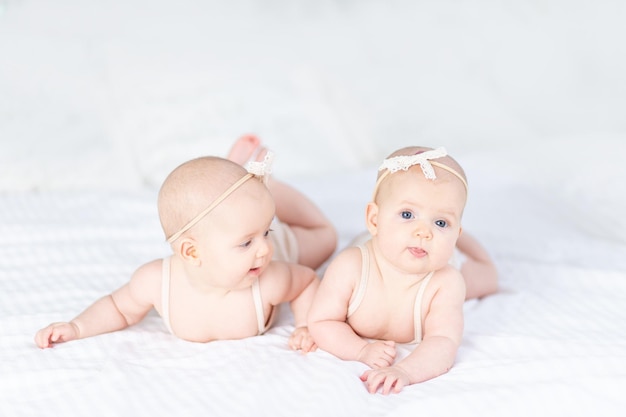 Deux filles jumelles nouveau-nées dans un costume en coton sur un lit blanc à la maison sont allongées sur le ventre et sourient en jouant les unes avec les autres