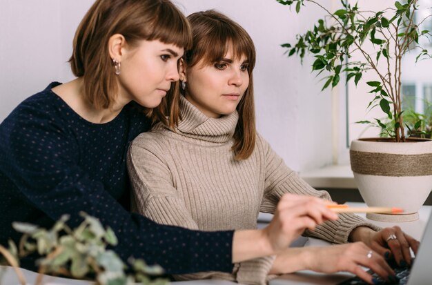 Deux filles jumelles travaillent côte à côte au bureau sur un ordinateur portable. Interaction des sœurs et des collègues au travail. La psychologie des relations de travail.