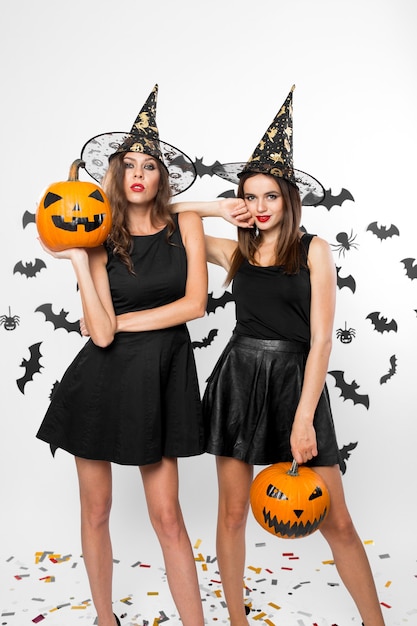 Deux filles magnifiques portant des robes noires et des chapeaux de sorcière tiennent des citrouilles d'halloween en arrière-plan avec des chauves-souris.