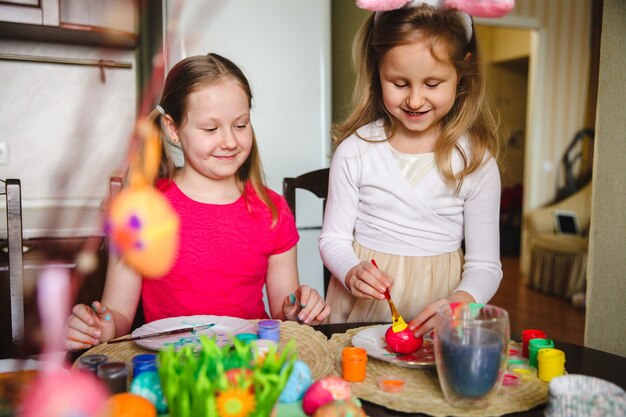 Deux filles à la maison dans la cuisine à la table peignent des œufs de Pâques avec de la peinture.