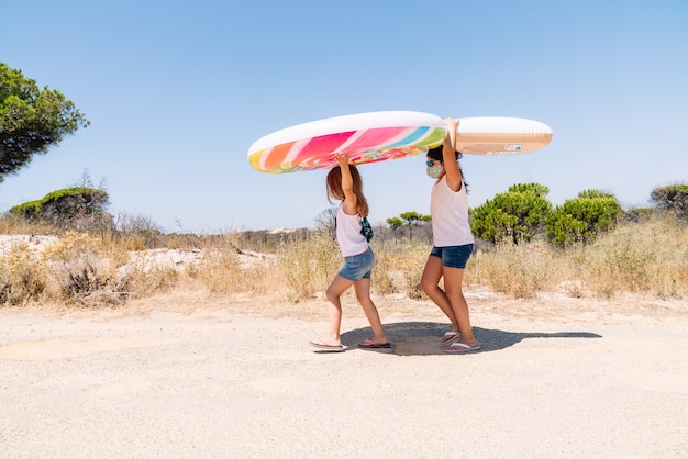 Deux filles avec un masque sur le visage et des lunettes se promenant autour d'un terrain de camping en pin avec une roue de matelas gonflable colorée sur la tête pour profiter de vacances au milieu du covid 19