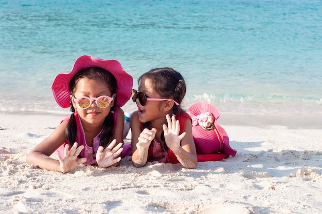 Photo deux filles mignonnes enfant asiatique portant chapeau rose et lunettes de soleil jouant avec du sable ensemble sur la plage