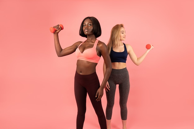 Deux filles minces internationales en vêtements de sport debout une par une, femme africaine et européenne tenant des haltères dans leurs mains et montrant leurs biceps