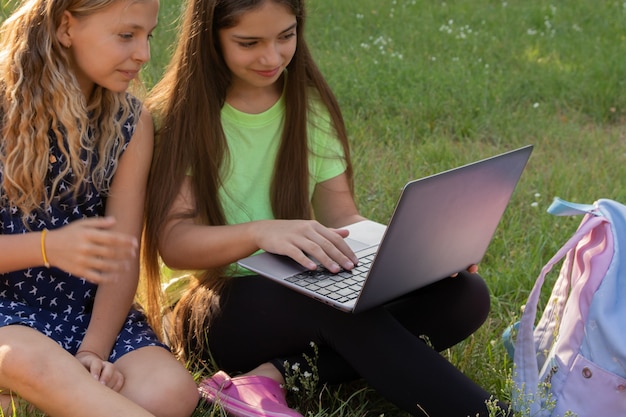 Deux filles avec un ordinateur portable et un sac à dos faisant leurs devoirs ou s'amusant assis sur l'herbe à l'extérieur