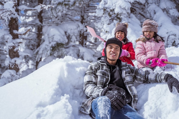 Photo deux filles et un père souriant assis sur un tas de neige profitent de leur temps ensemble en hiver enneigé