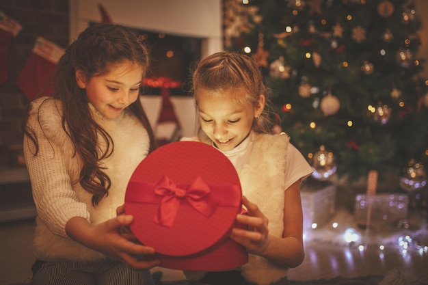 Deux filles souriantes sont assises par terre près du sapin de Noël. Surpris, ils regardent le cadeau dans une boîte rouge.