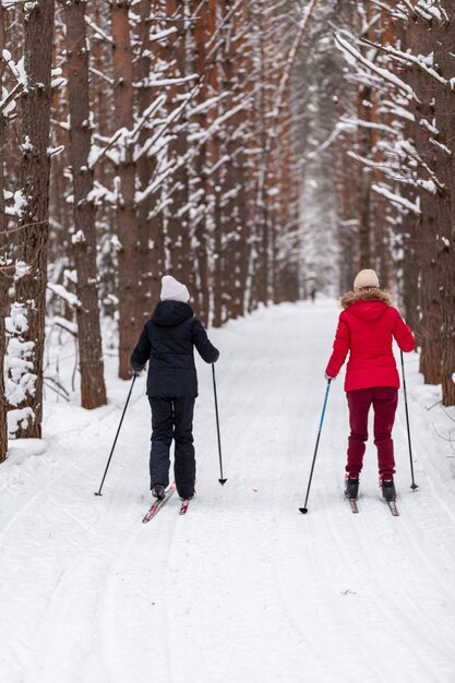 Deux filles vêtues d'une veste noire et rouge skient en hiver dans une forêt enneigée sur une piste de ski. Arbres dans une rangée.. Vue arrière. Skier dans une belle forêt enneigée dans le froid. belle nature hivernale