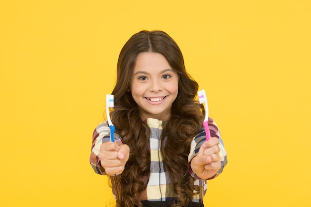 Deux fois plus rapide. Les cheveux longs mignons de fille tiennent le fond jaune de brosses à dents. Fille enfant détient deux brosses à dents. Le visage heureux d'enfant intelligent d'écolière d'enfant se soucie de l'hygiène. Concept de brosse à dents. Hygiène des dents.