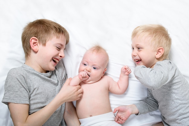 Deux frères aînés jouent et rient avec le plus jeune bébé dans un lit blanc, enfance heureuse, grande famille