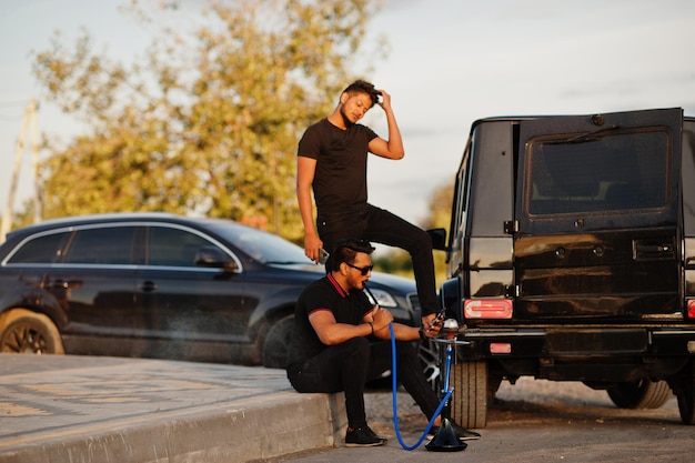 Deux frères asiatiques portent un homme tout noir posé près de voitures suv et fument le narguilé