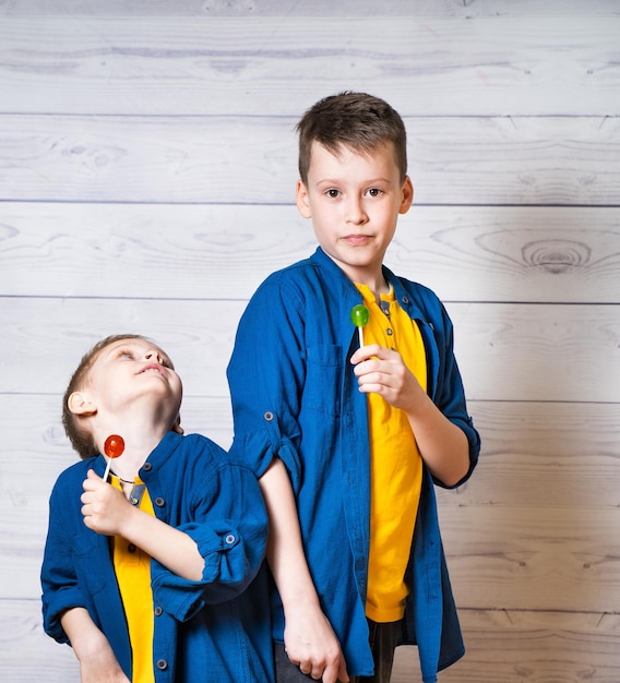 Deux frères en t-shirts jaunes et chemises bleues avec des sucettes colorées en studio Des garçons adorables s'amusent bien en léchant des sucettes sur un fond en bois clair